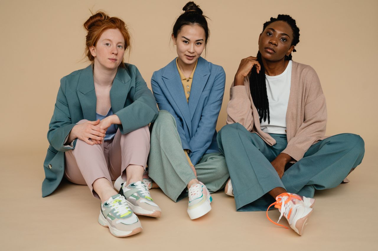 heros-img Three diverse women sitting in colorful suits against a beige backdrop, showcasing fashion and diversity.