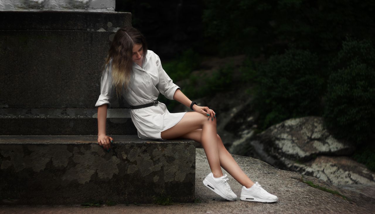 A young woman in a stylish white dress and sneakers sits gracefully on a stone bench outdoors, exuding elegance.